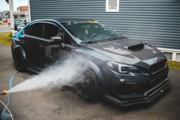 Nettoyage d'une voiture de sport sombre. Un jet d'eau est dirigé vers la roue avant, peut-être pour enlever mousse sur goudron vinaigre blanc.