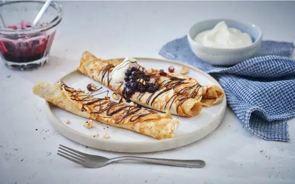 Matiga pannkakor rullade och toppade med chokladsås, blåbär och grädde. Serveras med en skål bärkompott och en blå rutig handduk.
