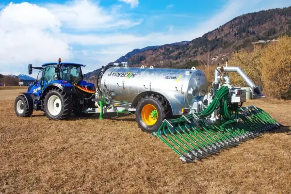 Niebieski traktor ciągnie przyczepę z cysterną do przewozu gnojowicy, wyposażoną w rozdzielacz z wieloma wężami do precyzyjnego nawożenia pól.