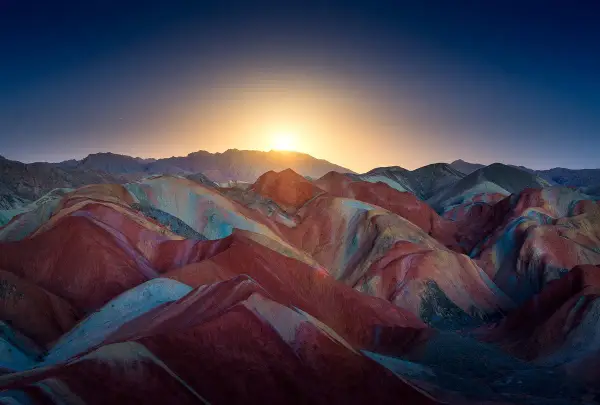 Bunte Schichten aus Rot, Gelb und Blau formen die **Regenbogenberge Chinas** unter einem tiefblauen Himmel mit aufgehender Sonne.
