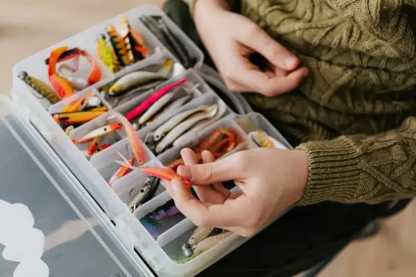 Ręka wybiera pomarańczową przynętę z pudełka pełnego różnorodnych akcesoriów do łowienia na żywca.