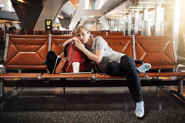 Zmęczona podróżniczka śpi na lotnisku, próbując pokonać jetlag. Obok niej plecak i kubek kawy.