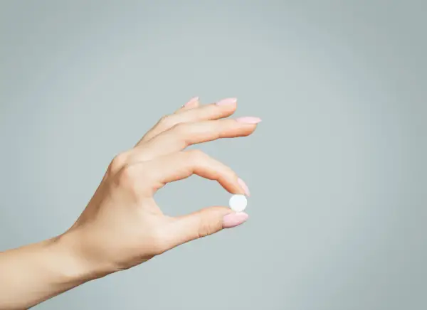 Dłoń z różowymi paznokciami trzyma białą tabletkę, być może zolpidem, na jasnoszarym tle.