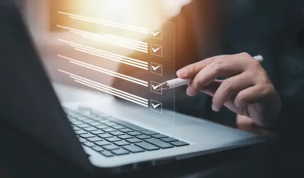 Ręka z długopisem zaznacza punkty na liście kontrolnej na ekranie laptopa. Proces ten może pomóc w ustaleniu, jak zmienić regulamin zakładowego funduszu świadczeń socjalnych.