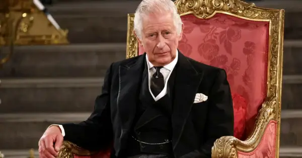 Król Karol w wieku siedemdziesięciu lat, w eleganckim czarnym garniturze, siedzi na ozdobnym tronie.