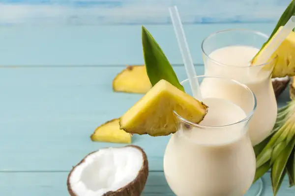 Dwa orzeźwiające koktajle Pina Colada, ozdobione kawałkami ananasa i liśćmi, na tle niebieskich desek. Idealny przepis na lato!