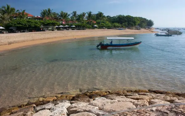 Spokojna zatoka w Sanur z łodzią "Water Walk" kołyszącą się na turkusowej wodzie. Piękna plaża z palmami i leżakami w tle.