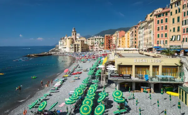 Plage animée à Camogli, Ligurie, avec des parasols verts et jaunes, des bâtiments colorés et la mer bleue.