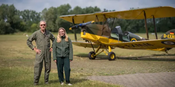 Mann in Fliegerkluft und junge Frau stehen neben gelbem Doppeldeckerflugzeug. Das Flugzeug erinnert an einen Tiger Moth.