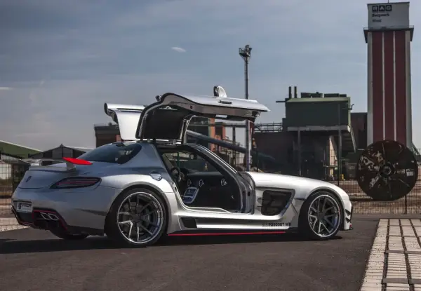 Srebrny SLS z otwartymi drzwiami typu gullwing, z czerwonym akcentem na spojlerze, zaparkowany przed industrialnym budynkiem.