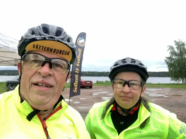 Två cyklister redo för Vätternrundan. Mannen har en keps med texten "Vätternrundan". Bakom dem syns en sjö och en flagga för Sollers IF Fotboll.