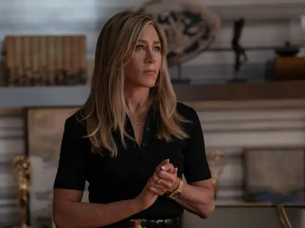 Jennifer Aniston zamyślona, w czarnej bluzce, z dłońmi złożonymi na piersi. W tle widać elementy wystroju wnętrza, może nawiązujące do jej życia, jakby myślała o swoich przyszłych dzieciach.