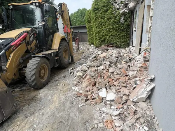 Koparka pracuje przy rozbiórce domu. Gruz i cegły leżą na ziemi, szacowany koszt rozbiórki domu jest wysoki.