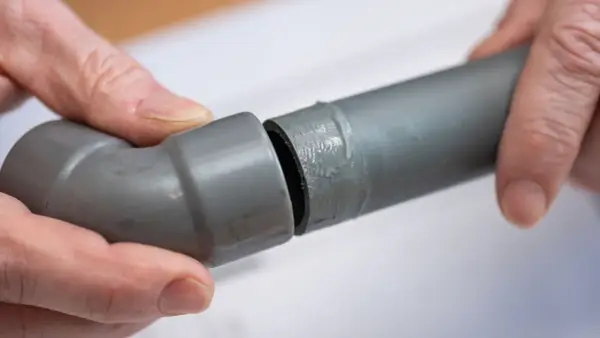 Mains de personne assemblant deux tuyaux gris en **pvc renforcé**, l'un avec un coude, prêts pour une installation solide.