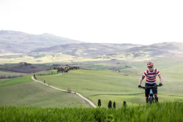 Ciclista in un paesaggio collinare della Toscana, con un sentiero tortuoso che serpeggia tra campi verdi.