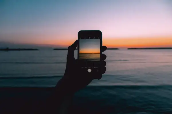 Dłoń trzyma smartfon, na ekranie którego widać piękny zachód słońca nad morzem. Idealny kadr, by nauczyć się, jak fotografować zachód słońca.