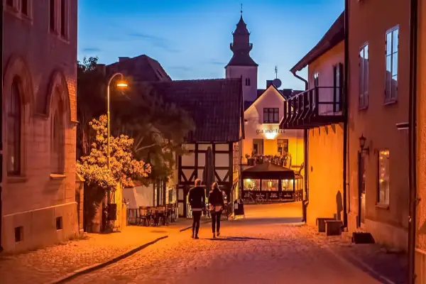 Två personer promenerar på en kullerstensgata i Båstad under Stockholmsveckan. Kvällen är ljum, och husen längs gatan lyser varmt.