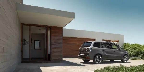 Szary minivan Citroën C4 Picasso zaparkowany przed nowoczesnym domem z drewnianą elewacją.