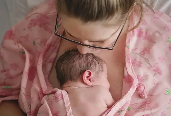 Mama całuje główkę noworodka, otulając go w różowy materiał. To piękny moment kangurowania, budowania więzi.