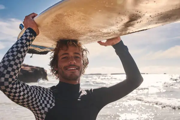 Lächelnder Surfer mit lockiger surfer frisur trägt Neoprenanzug und hält Surfbrett über dem Kopf.