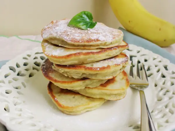 Stos puszystych, trzyskładnikowych placków bananowych, posypanych cukrem pudrem i udekorowanych listkiem, obok dojrzałego banana.