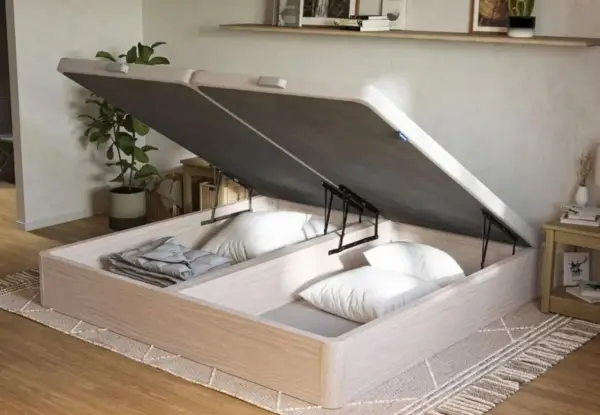 Un canapé de madera clara con la tapa abierta, revelando un gran espacio de almacenamiento para ropa de cama y almohadas.