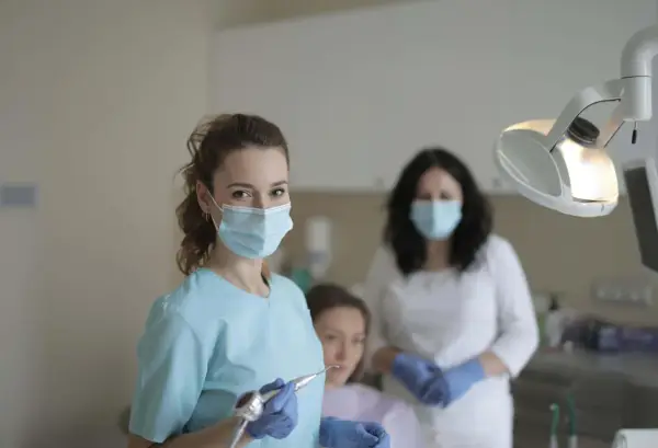 Uśmiechnięta studentka stomatologii w masce i rękawiczkach trzyma wiertło dentystyczne. W tle inne osoby w gabinecie.