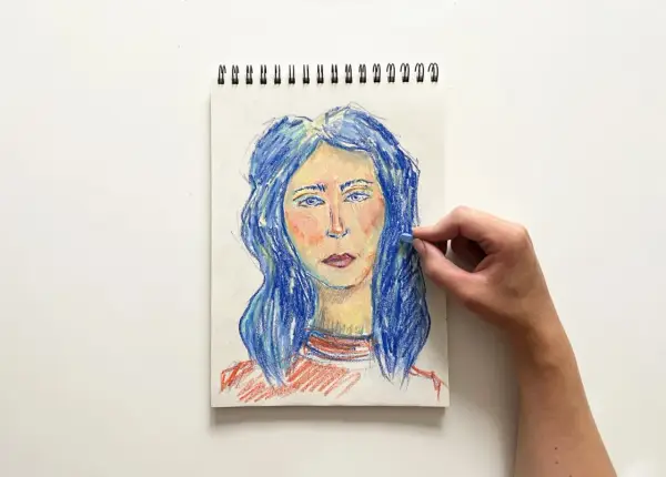 Portret kobiety z niebieskimi włosami, tworzony pastelami. Ręka dodaje ostatnie pociągnięcia, tworząc efekt dragana.
