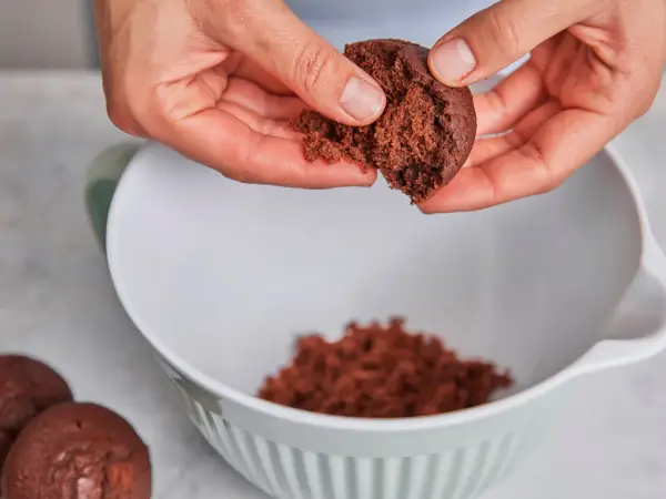 Hände zerbröseln einen Schokoladenkuchen für vegane Desserts. Ein Teil landet in einer weißen Schüssel.