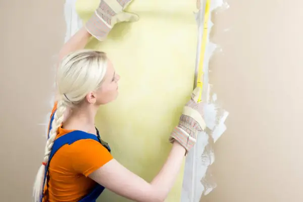 Une femme en salopette bleue et t-shirt orange mesure un mur pour le détapisser puis peindre.