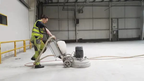 Pracownik w kamizelce odblaskowej wykonuje szlifowanie betonu za pomocą maszyny.