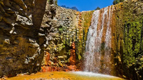 Ein Wasserfall stürzt über moosbewachsene, orangefarbene Felsen. Die Felsen sind von der Sonne beschienen, und das Wasser sammelt sich in einem goldenen Becken. Ein Stück Natur auf La Palma.