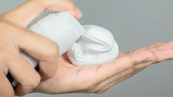 Mano aplicando gel o espuma de afeitar de un envase blanco a la palma.