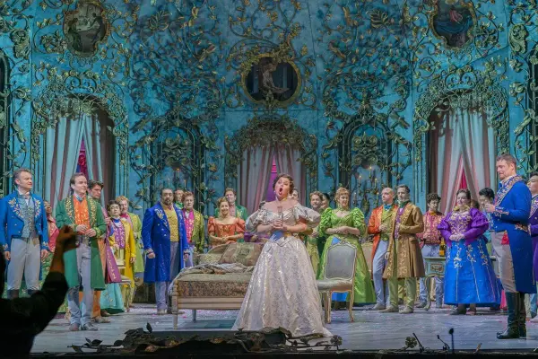 Primadonna w lśniącej sukni śpiewa na scenie operowej, otoczona przez barwnie ubranych artystów.