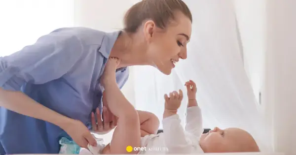Uśmiechnięta mama pochyla się nad dzieckiem, które leży na plecach. Wykonuje szerokie pieluchowanie, delikatnie unosząc nóżki malucha.