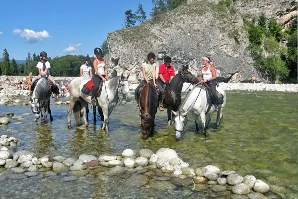 Konni jeźdźcy na koniach w rzece, w tle skaliste zbocze i plaża z ludźmi. To widok z Bukowiny Tatrzańskiej, jakby rynek atrakcji.