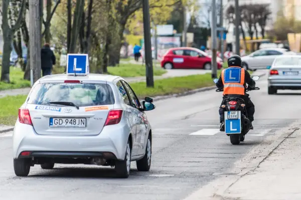 Srebrny samochód z literą "L" na dachu i motocyklista w kamizelce odblaskowej, obaj z literą "L" z tyłu, poruszają się po drodze. To kategoria a nauki jazdy.