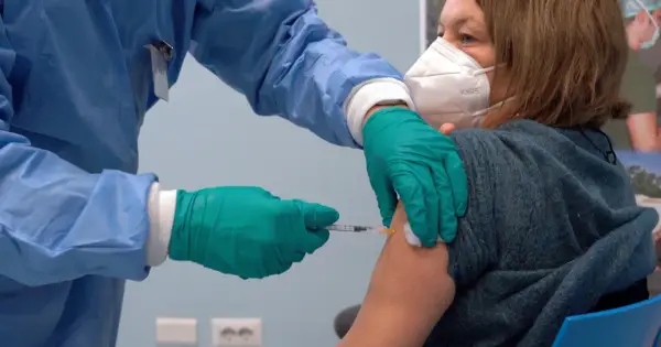 Osoba w niebieskim fartuchu i zielonych rękawiczkach podaje zastrzyk. Kobieta w masce KN95 otrzymuje trzecią dawkę szczepionki.