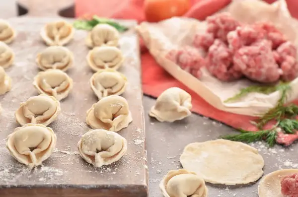 Gotowe do lepienia pierogi z mięsem mielonym surowym. Na desce ułożone pierożki, obok surowe mięso i ciasto.