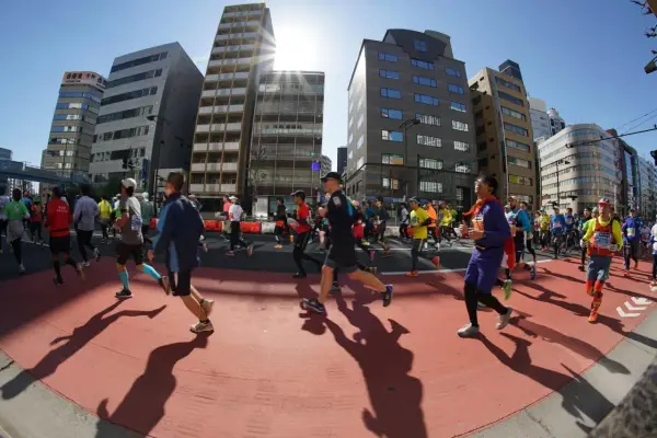 Atleti corrono nella maratona di Tokyo, circondati da grattacieli moderni sotto un sole splendente.