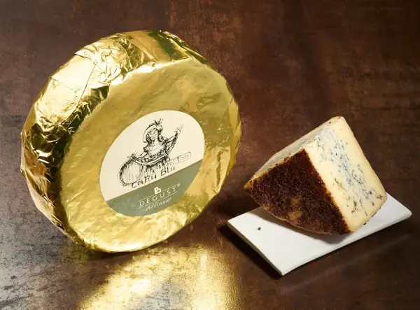 Formaggio blu con crosta scura, accanto a una forma intera avvolta in carta dorata. Questo cacio cos'è? Un'eccellenza da gustare.