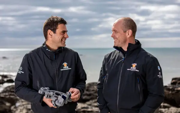 Damien Guillou et son collègue souriants sur la plage, l'un tenant un sextant, prêts pour une nouvelle aventure maritime.