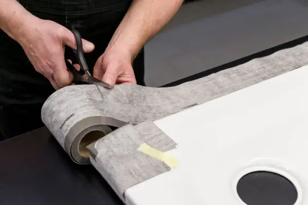 Hände schneiden Dichtungsband zu, um eine Duschwanne abzudichten. Das Material ist auf einer Rolle und wird präzise zugeschnitten.