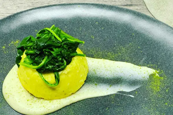 Flan di pecorino giallo, sormontato da spinaci freschi, su una crema bianca e polvere verde su piatto scuro.