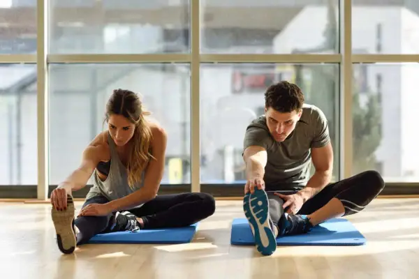 Para ćwiczy stretching. Kobieta i mężczyzna siedzą na macie, wyciągając nogi i dotykając palców u stóp.