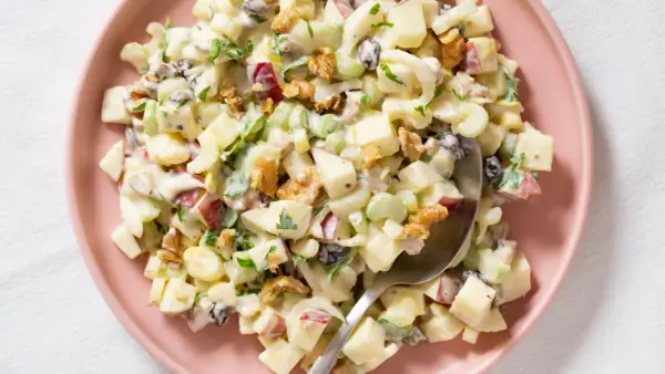 Ensalada Waldorf cremosa con trozos de manzana, apio, nueces y pasas, servida en un plato rosa.