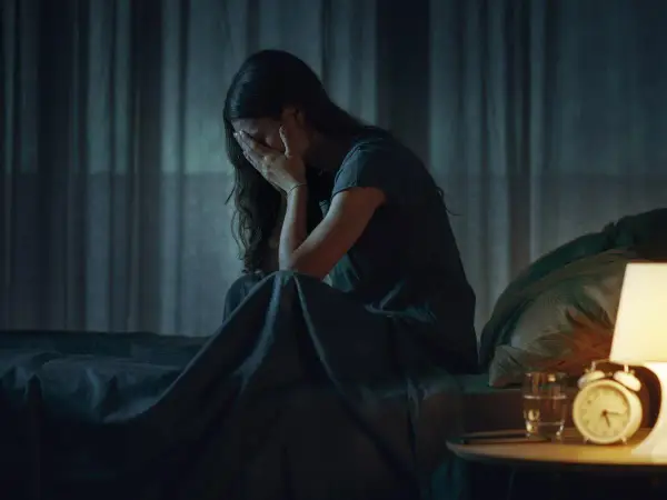 Mujer sentada en la cama, cubriéndose la cara con las manos, en plena vigilia del sueño. La noche es oscura, solo iluminada por una lámpara.