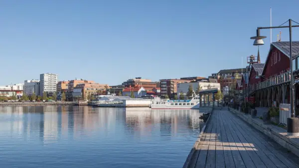 En vacker dag att besöka Luleå. En träspång leder längs vattnet förbi röda byggnader och en vit båt.