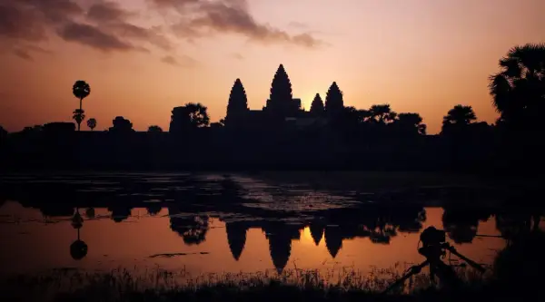 Zachód słońca nad Angkor Wat, idealne tło dla niezapomnianych kambodżańskich wakacji. Widok odbija się w wodzie, a na pierwszym planie statyw z aparatem.