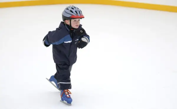 Bambino con casco e tuta blu pratica il pattinaggio sul ghiaccio, con i pattini azzurri e lacci arancioni.